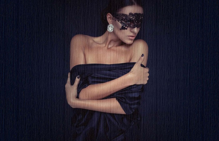 Mulher com máscara preta e vestido preto, abraçada a si e com ar sensual na casa das máscaras sexy.